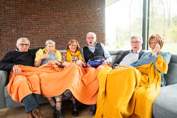 This lively image showcases a group of seniors collectively reacting while seated on a couch, absorbed in watching a riveting television program. Their expressions are a mix of surprise, concern, and