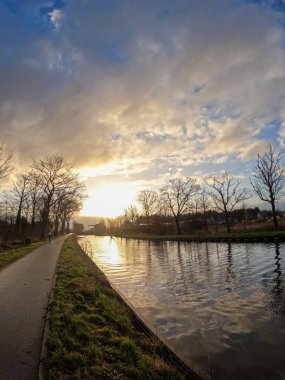 Nefes kesen güzel bir gün batımı güzel bir şekilde ağaçlarla kaplı sakin bir kanala ve yürüyen bir yola yansıyor.