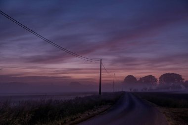 Rengarenk şafak vakti gökyüzünün altında dolambaçlı bir yolu olan sakin bir kır manzarası. Sabah sisiyle kaplanmış.
