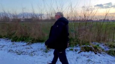 Bir adam, açık arazilerle çevrili, karlı bir yolda sakin bir şekilde yürür. Arka planda güzel bir günbatımı vardır.