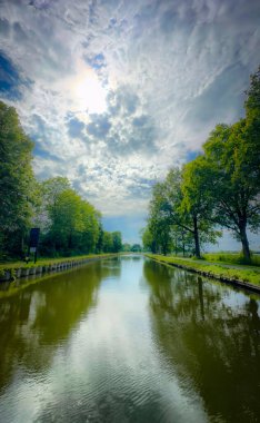 Huzurlu ve sakin bir nehir manzarası sergileniyor çarpıcı yansımalar ve dramatik bulutlar havada süzülüyor