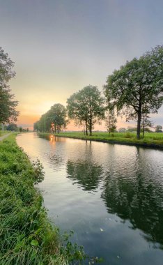 Sakin bir nehir serin bir atmosferdeki yemyeşil ağaçlar tarafından çerçevelenmiş çarpıcı bir günbatımının güzelliğini yakalar.