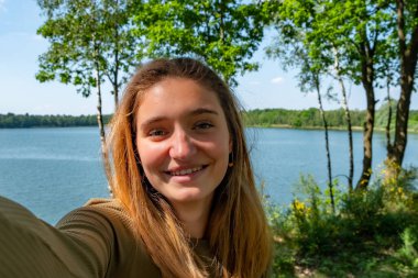 Neşeli genç bir kadın, güzel yeşillikler ve ağaçlarla çevrili sakin bir gölün kenarında mutlu bir selfie çeker.