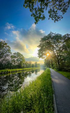 Huzurlu bir nehir manzarası. Sabahın erken saatlerinde aydınlanan huzurlu bir yolu çevreleyen yemyeşil bir yer.
