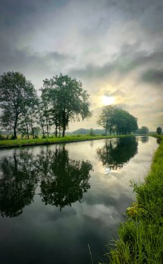 Ağaçları ve bulutları yansıtan huzurlu bir nehir manzarası, barışçıl doğanın güzelliğini yakalıyor.