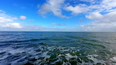 Yumuşak okyanus dalgaları, mavi gökyüzünün altında tüylü bulutlarla sahili okşar ve sakin bir sahil manzarası yaratır.