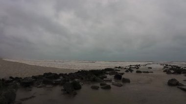 Resimli bir sahil sahnesi, bulutlu bir gökyüzünün altındaki engebeli kayalara güçlü bir şekilde çarpan dalgaları gözler önüne seriyor.