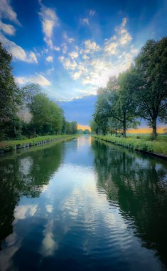 Canlı ağaçlar ve dramatik bulutlarla kaplı sersemletici yansımaları gösteren huzurlu ve huzurlu bir nehir manzarası.