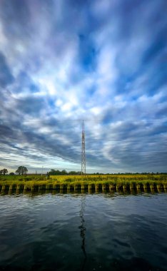 Güzel ve sakin bir su manzarası, dinamik ve canlı bulutların üzerinde yükselen yüksek bir iletişim kulesine sahiptir.
