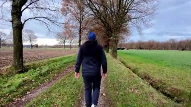 Bir insan verimli ağaçlar ve açık alanlarla çevrili manzaralı bir yolda rahatça yürüyor, doğanın tadını çıkarıyor.