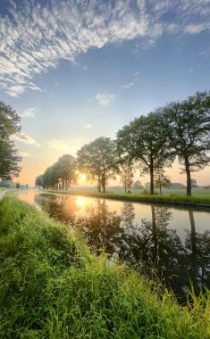 Gün batımında yavaşça akan sakin bir nehir, sudaki canlı renkleri yansıtıyor ve yemyeşil ile çevrili.