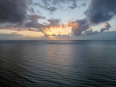 Gün batımında gökyüzünün sıcak renklerini yansıtan bulutlarla birlikte engin okyanusun güzel, sakin bir görüntüsü.