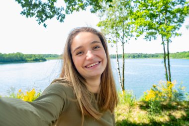 Neşeli genç bir kadın, dingin bir gölün kenarında, canlı yeşilliklerle çevrili, huzurlu bir sahne yaratarak eğleniyor.