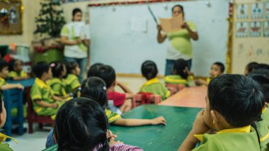 Üniforma giymiş anaokulu çocukları okulda oturan ve bayan öğretmenlerin derslerini dinliyorlar.