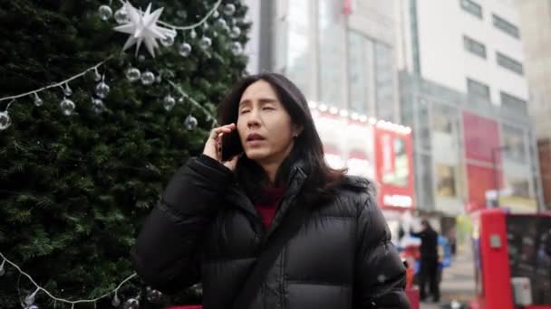 30代の韓国人男性が ソウル 明洞で寒くて雪の多いクリスマスツリーの前でスマートフォンに電話をかけます — ストック動画