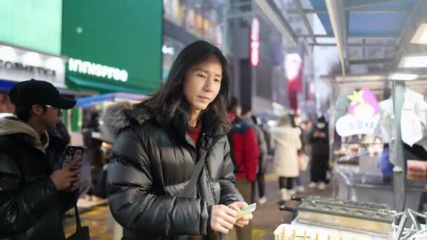 韩国首尔明东的一个寒冷的雪夜 一个30多岁的长发韩国男人在夜市里购买和吃小吃 — 图库视频影像