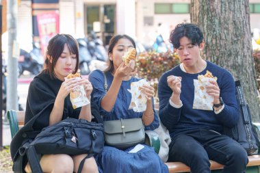 Tayvanlı ve Asyalı üç üniversite öğrencisi Tayvan 'ın Taipei şehrindeki Da' an Bölgesi 'ndeki bir bankta sokak yemeği yiyorlar.