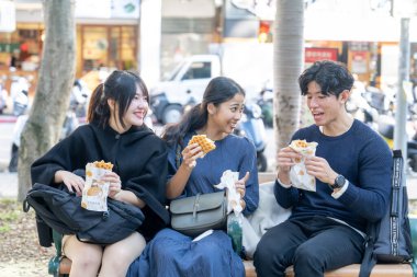 Tayvanlı ve Asyalı üç üniversite öğrencisi Tayvan 'ın Taipei şehrindeki Da' an Bölgesi 'ndeki bir bankta sokak yemeği yiyorlar.