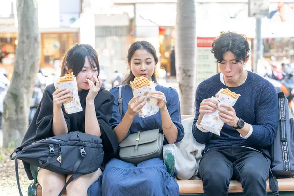Three Taiwanese and Asian college students eat street food waffles on a bench around the university in Da\'an District, Taipei City, Taiwan