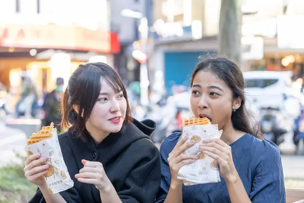 Three Taiwanese and Asian college students eat street food waffles on a bench around the university in Da\'an District, Taipei City, Taiwan
