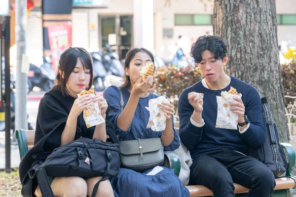 Three Taiwanese and Asian college students eat street food waffles on a bench around the university in Da'an District, Taipei City, Taiwan