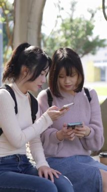 Tayvan, Taipei 'de bir üniversite kampüsünde oturan ve mutlu bir şekilde konuşan iki genç Tayvanlı kız öğrencinin yavaş çekimde dikey videosu.