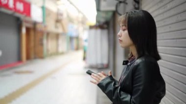 Okinawa bölgesinden 20 'li yaşlarda, Okinawa' nın Naha şehrindeki Kokusai-dori Caddesi yakınlarındaki bir alışveriş sokağında kışın giydiği kıyafetlerle duran ve akıllı telefonunu kullanan genç bir Japon kadın.