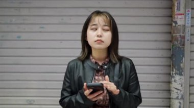 Okinawa bölgesinden 20 'li yaşlarda, Okinawa' nın Naha şehrindeki Kokusai-dori Caddesi yakınlarındaki bir alışveriş sokağında kışın giydiği kıyafetlerle duran ve akıllı telefonunu kullanan genç bir Japon kadın.