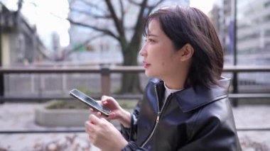 20 'li yaşlarda Japon bir kadın kışın Tokyo, Shinagawa' daki Gotanda İstasyonu 'nda halka açık bir yolda akıllı telefon kullanıyor.