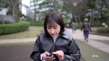 Japon bir kadının kışın Gotanda İstasyonu, Shinagawa-ku, Tokyo yakınlarındaki bir parkta oturup akıllı telefonunu kullanmasını gösteren yavaş çekim videosu.