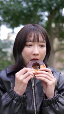 20 'li yaşlarda genç bir Japon kadının Gotanda, Shinagawa-ku, Tokyo yakınlarındaki bir parkta oturup donut yerken çekilmiş dikey videosu.