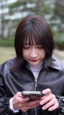 20 'li yaşlarda genç bir Japon kadının Gotanda İstasyonu, Shinagawa-ku, Tokyo yakınlarındaki bir parkta oturmuş akıllı telefonunu kullanışının dikey videosu.
