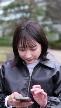 20 'li yaşlarda genç bir Japon kadının Gotanda İstasyonu, Shinagawa-ku, Tokyo yakınlarındaki bir parkta oturmuş akıllı telefonunu kullanışının dikey videosu.