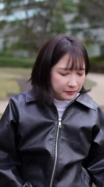 20 'li yaşlarda genç bir Japon kadının Gotanda İstasyonu, Shinagawa-ku, Tokyo yakınlarındaki bir parkta oturmuş akıllı telefonunu kullanışının dikey videosu.