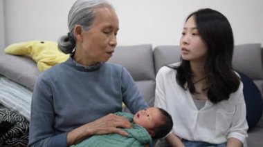 70 'li yaşlarda Tayvanlı bir büyükanne, elinde beyaz kostümlü 8 günlük bir bebek varken 30' lu yaşlarda Tayvanlı bir kadının kızıyla evdeki koltukta konuşuyor..