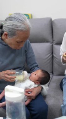 70 'li yaşlarda Tayvanlı bir büyükannenin ve 30' lu yaşlarda bir annenin beyaz kostümlü 8 günlük bir bebeği kucağına alıp kanepede süt verişinin yavaş çekimde dikey videosu.