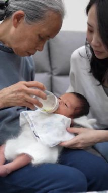 70 'li yaşlarda Tayvanlı bir büyükannenin ve 30' lu yaşlarda bir annenin beyaz kostümlü 8 günlük bir bebeği kucağına alıp kanepede süt verişinin yavaş çekimde dikey videosu.