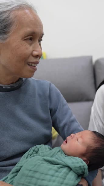 70代の台湾人の祖母と30代の母親のスローモーションの垂直ビデオは 白い衣装で8日間の赤ちゃんを抱えてソファーで遊んでいます — ストック動画