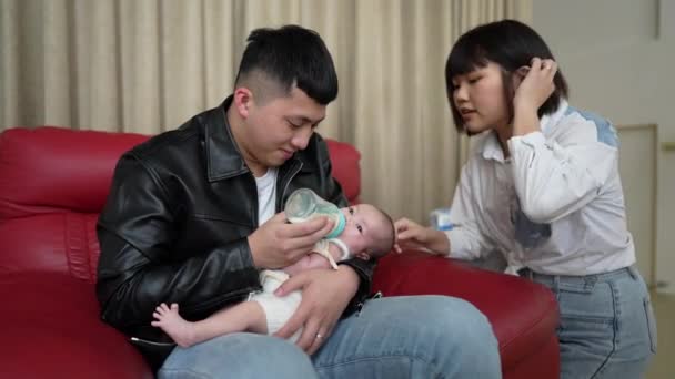 一位20多岁的台湾籍母亲和一位父亲坐在一张红色的沙发上 正在给一位一个月大的女婴喂奶 — 图库视频影像