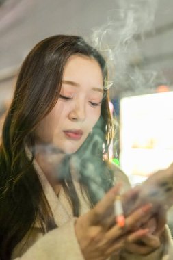 20 'li yaşlarda bir Çinli kadın, Saitama, Saitama' da Omiya Ward 'daki istasyonun önündeki sigara içme alanında akıllı telefonunu kullanırken sigara içiyor.