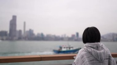 Hong Kong 'un Causeway Körfezi' nde genç bir kadın denizin kenarında dikilip, düşünceli bir şekilde şehrin ufuk çizgisine bakıyor..