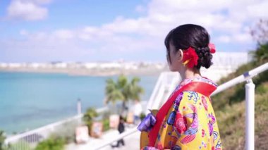 Geleneksel Ryusou giyimli yirmili yaşlarda genç bir kadın Okinawa 'da bir sahil kasabasının merdivenlerinde geziniyor ve mekanın kültürel zarafetini ve manzarasının güzelliğini vurguluyor..