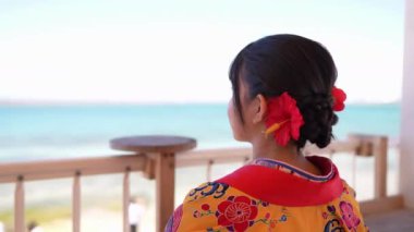 Geleneksel Ryusou giyen yirmili yaşlarında genç bir kadın Okinawa 'da deniz kenarında oturup denize bakıyor. Sahne, Okinawa 'nın dingin güzelliğini ve kültürel mirasını vurguluyor..