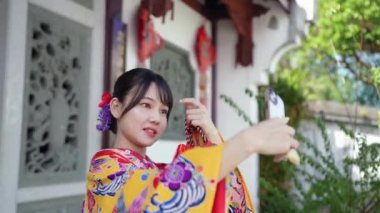 Geleneksel Ryusou giyen yirmili yaşlarda genç bir kadın Okinawa 'da Çin tarzı bir bahçede akıllı telefonuyla selfie çekiyor. Bu sahne kültürel miras ve modern teknolojinin karışımını vurguluyor.