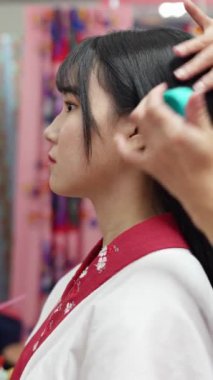 Geleneksel giysiler giyen yirmili yaşlarında genç bir kadın saçını ve makyajını Okinawa 'da içeride yaptırıyor. Bu dikey yavaş çekim videosu kültürel miras ve modern güzelliğin karışımını vurgular.