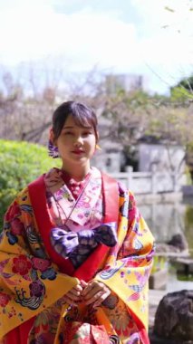 Geleneksel kıyafetler giyen yirmili yaşlarında genç bir kadın Okinawa 'da Çin tarzı bir bahçede yürüyor. Bu dikey yavaş çekim videosu kültürel miras ve doğal güzelliğin karışımını vurguluyor..