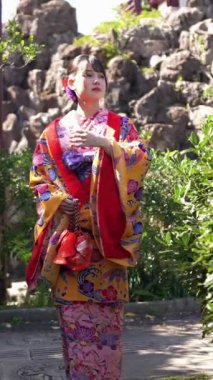 Geleneksel kıyafetler giyen yirmili yaşlarında genç bir kadın Okinawa 'da Çin tarzı bir bahçede yürüyor. Bu dikey yavaş çekim videosu kültürel miras ve doğal güzelliğin karışımını vurguluyor..