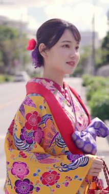 Geleneksel kıyafetler giyen yirmili yaşlarında genç bir kadın Okinawa 'da bir kasabada yürüyor. Bu dikey yavaş çekim videosu kültürel miras ve kentsel yaşamın karışımını vurguluyor.