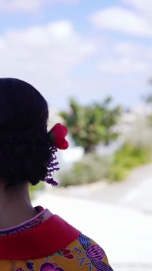 Geleneksel kıyafetler giyen yirmili yaşlarında genç bir kadın elinde plastik bir fincan kahveyle deniz kenarında yürüyor. Bu dikey yavaş çekim videosu kültürel miras ve modern yaşamın karışımını vurguluyor..