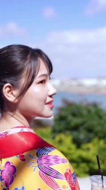 Geleneksel kıyafetler giyen yirmili yaşlarında genç bir kadın elinde plastik bir fincan kahveyle deniz kenarında yürüyor. Bu dikey yavaş çekim videosu kültürel miras ve modern yaşamın karışımını vurguluyor..